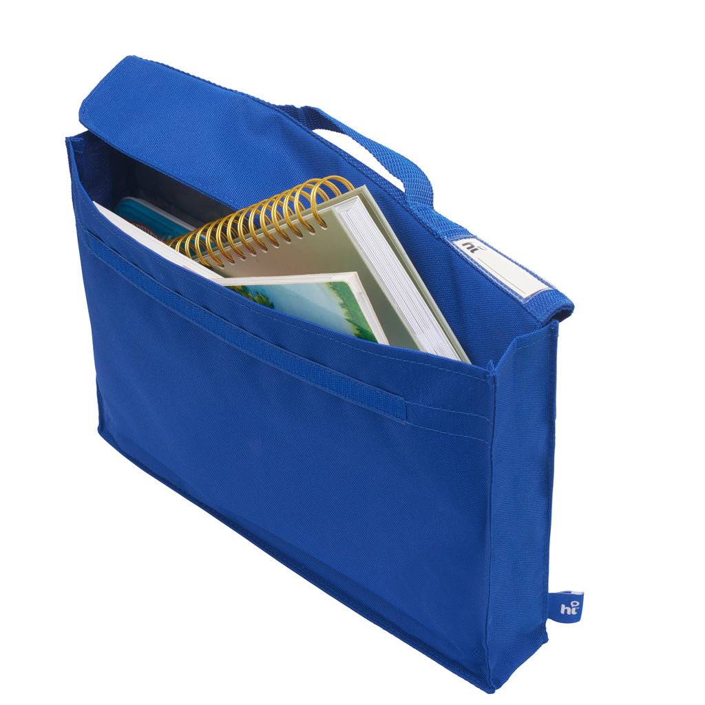 Front angle down view of the Harlequin Premier Library Bag in royal blue, showing the velcro closure, top handle and name tag. The bag has been packed with school books and a tin of coloured pencils.