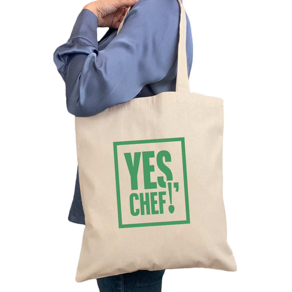 Women standing with calico bag over her shoulder, side view. Natural colours calico bag with green type design - Yes Chef!
