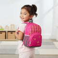 Child wearing a pink backpack with plaid pattern and butterflies on the pocket, she is standing looking after her shoulders indoors