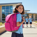 Girl with a pink backpack standing in front of a school building with other students in the background.